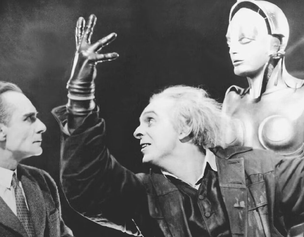 black and white film still with two men, one with white hair and hand raised, in front of a mannequin