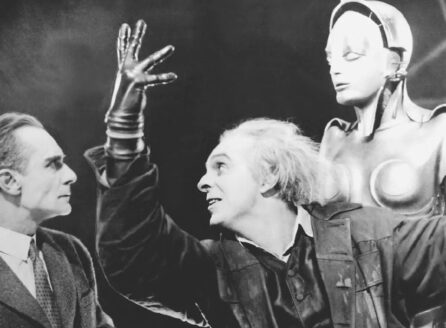 black and white film still with two men, one with white hair and hand raised, in front of a mannequin