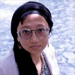 Shireen Hamza wearing glasses, hijab, white blazer, earrings, and patterned shirt