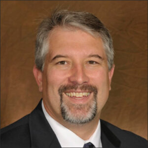 Erik G. Herbert smiling, wearing a suit, over bronze studio background.