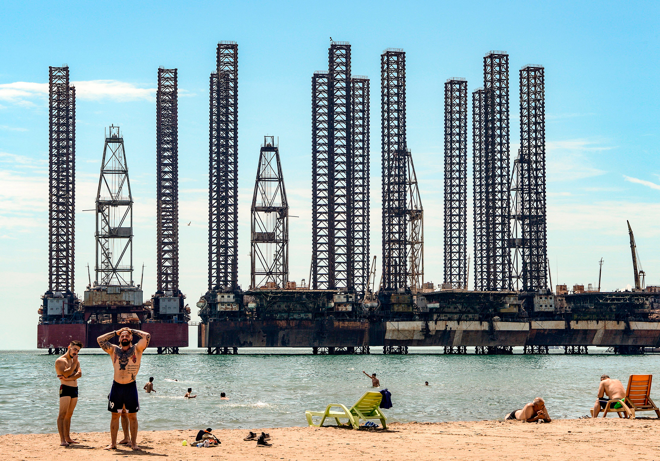 Photo of men on a beach with a line of oil rigs looming over them