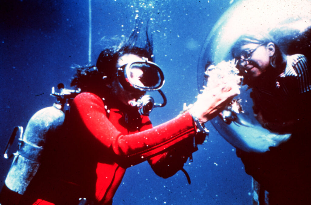 Scuba diver in water showing a sample to a woman behind curved glass