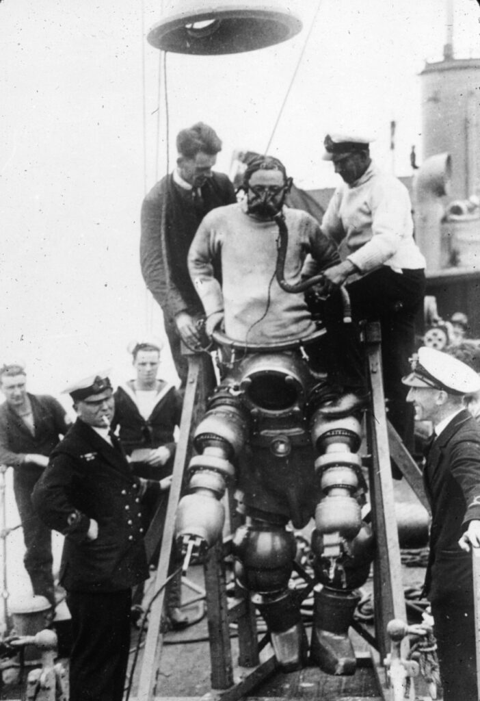 Old photo of man entering complicated diving suit with help of others