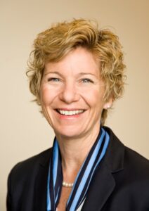 Sue Desmond-Hellmann over a tan background, smiling, wearing navy blue jacket and silk striped scarf