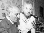 Black and white photo of a seated older man and standing older woman in front of a microscope