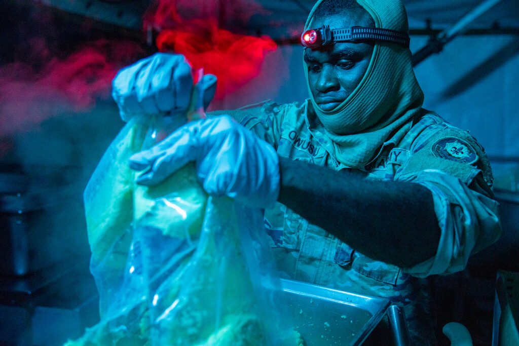 Color photo of an infantryman wearing battle fatigues and a headlamp opening a large bag of shelf-stable eggs