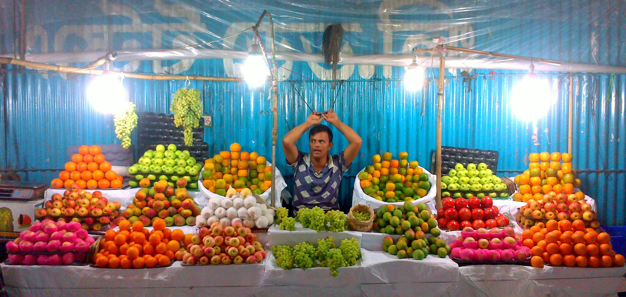 Man sitting at a brightly lit outdoor fruit stand at night