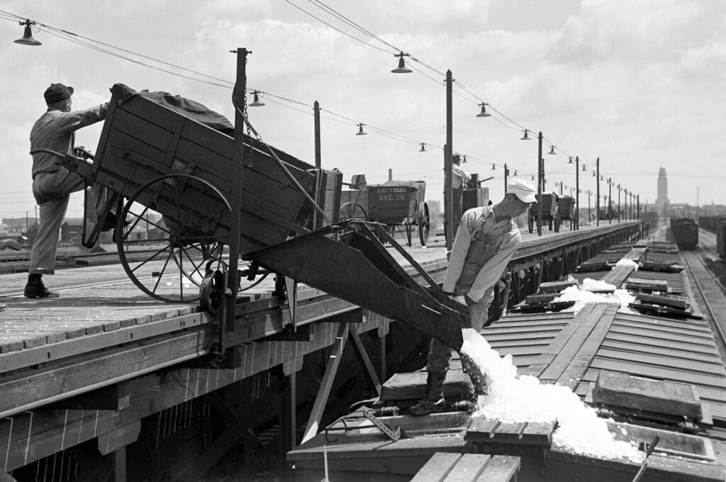 Black and white photo of two workers pouring ice into the top of a train car from a wagon on an elevated platform