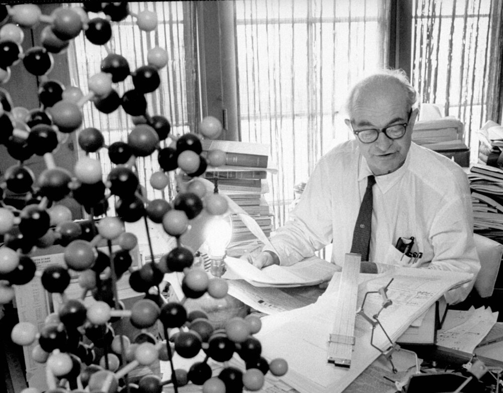 Older man sitting at a desk stacked with books, a molecular model in the foreground