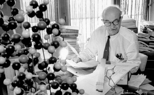 Older man sitting at a desk stacked with books, a molecular model in the foreground