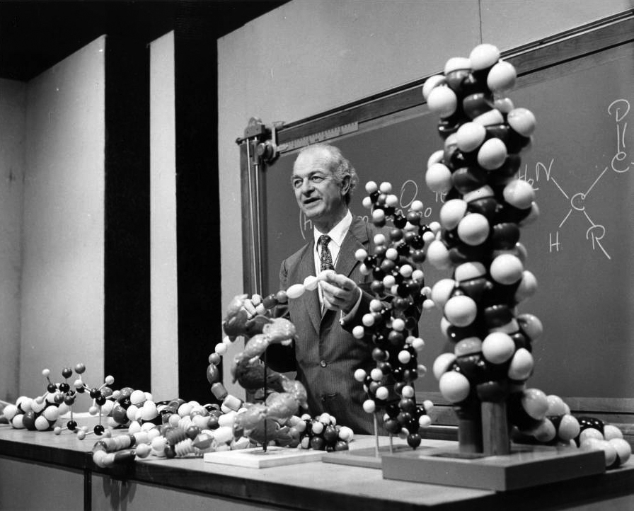 Older man in the front of a classroom auditorium, standing with models on a demonstration table