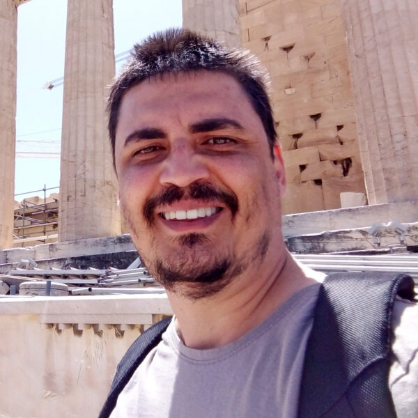 man in t-shirt and backpack, smiling in front of Ancient Greek columns