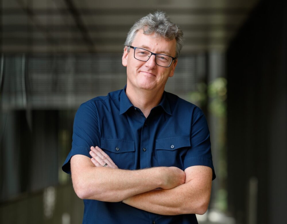 man with glasses and dark blue short-sleeved shirt, folded arms, in a hallway