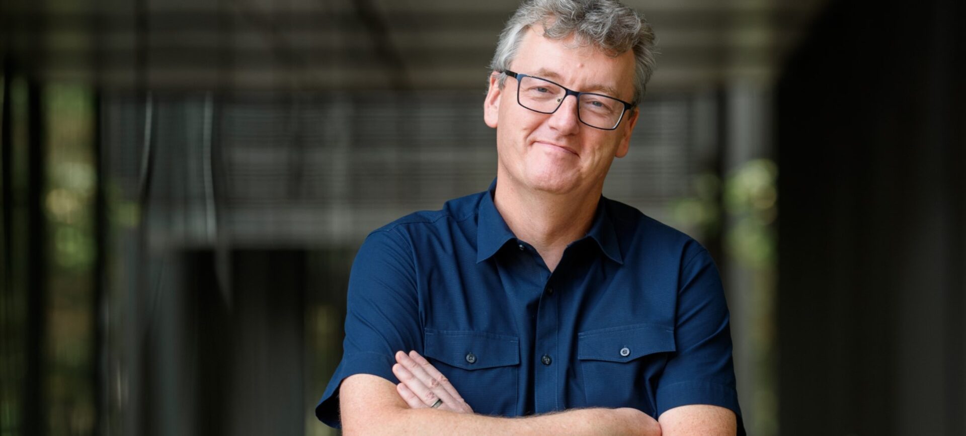 man with glasses and dark blue short-sleeved shirt, folded arms, in a hallway