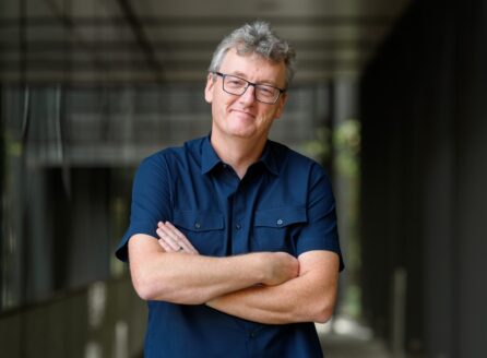 man with glasses and dark blue short-sleeved shirt, folded arms, in a hallway