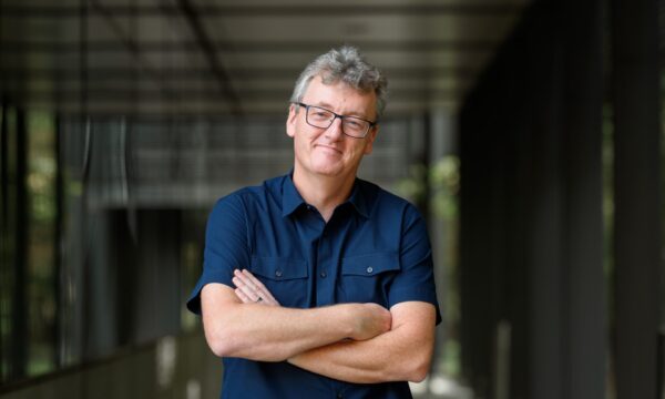man with glasses and dark blue short-sleeved shirt, folded arms, in a hallway