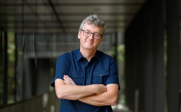 man with glasses and dark blue short-sleeved shirt, folded arms, in a hallway