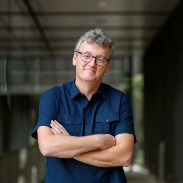 man with glasses and dark blue short-sleeved shirt, folded arms, in a hallway