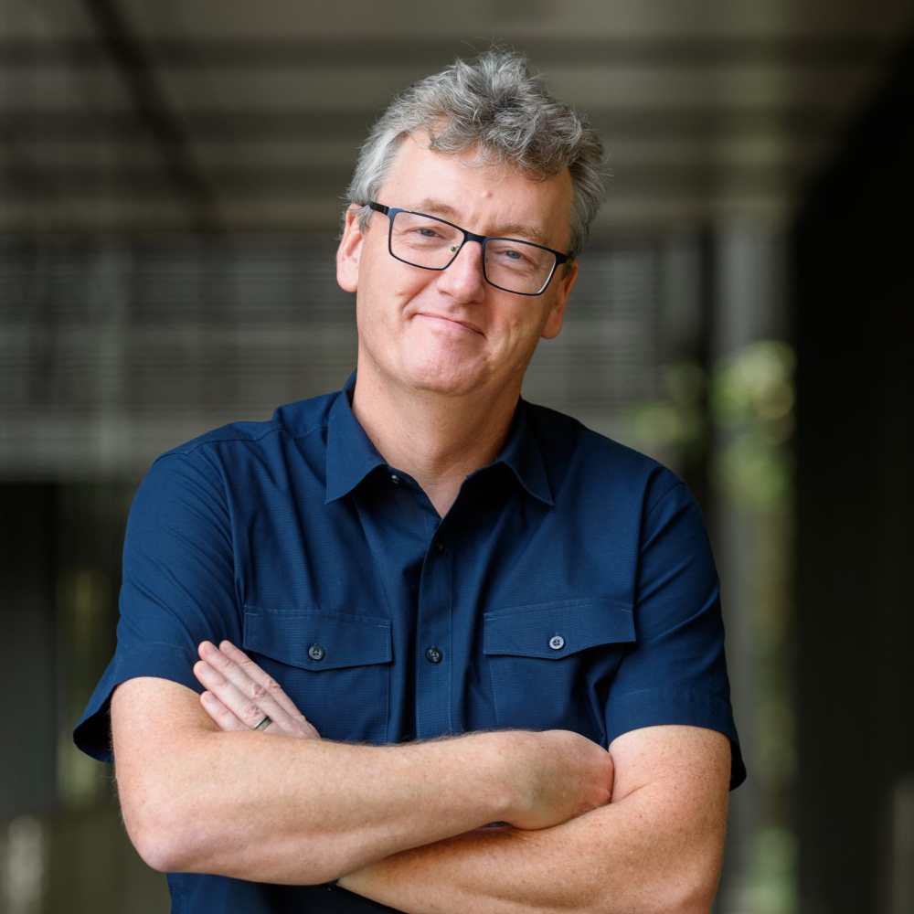 man with glasses and dark blue short-sleeved shirt, folded arms, in a hallway