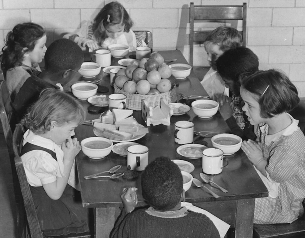 Black and white photo of school children sitting around a table eating lunch