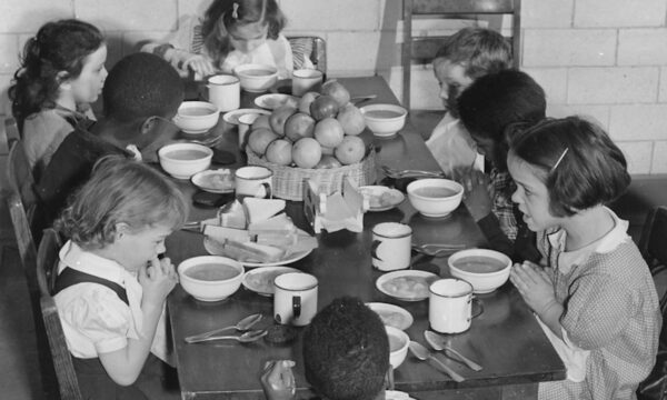 Black and white photo of school children sitting around a table eating lunch