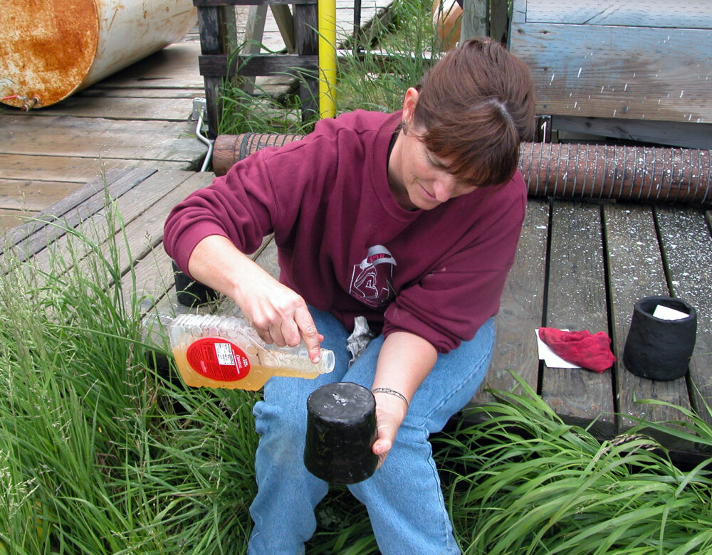 Seated woman pouring liquid from a plastic container onto an upturned pot