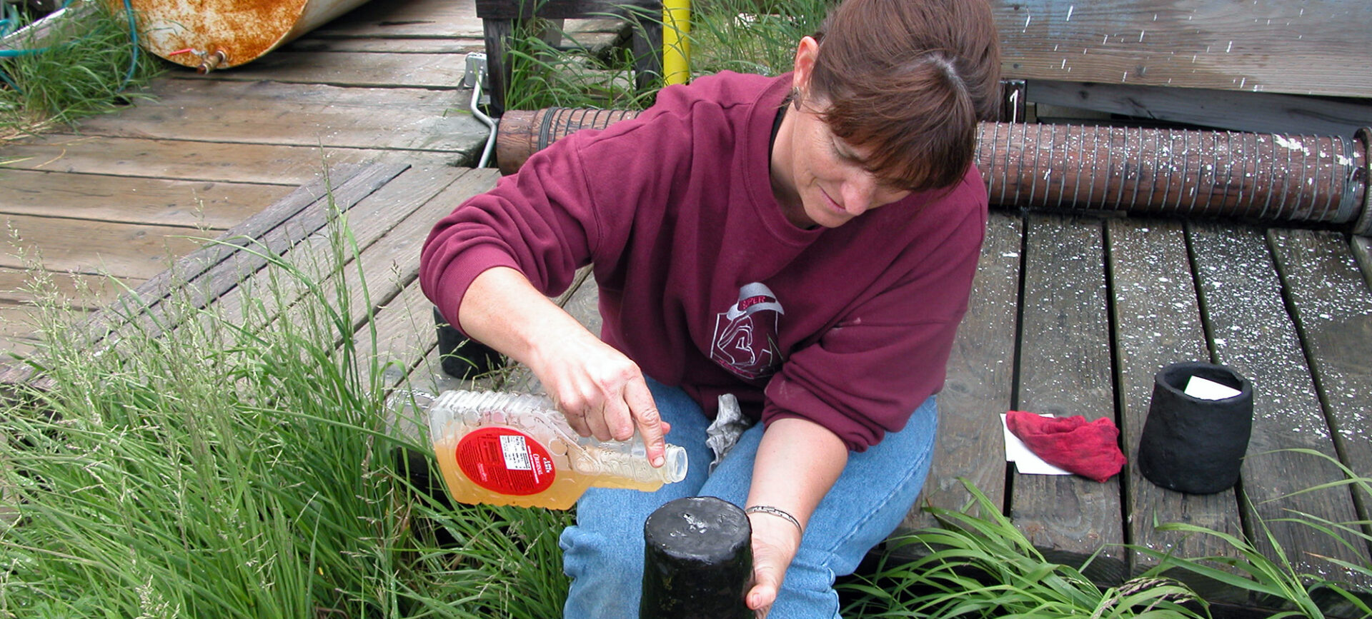 Seated woman pouring liquid from a plastic container onto an upturned pot
