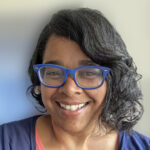 Jacqueline, smiling, wearing blue glasses and navy blue/coral layered shirts