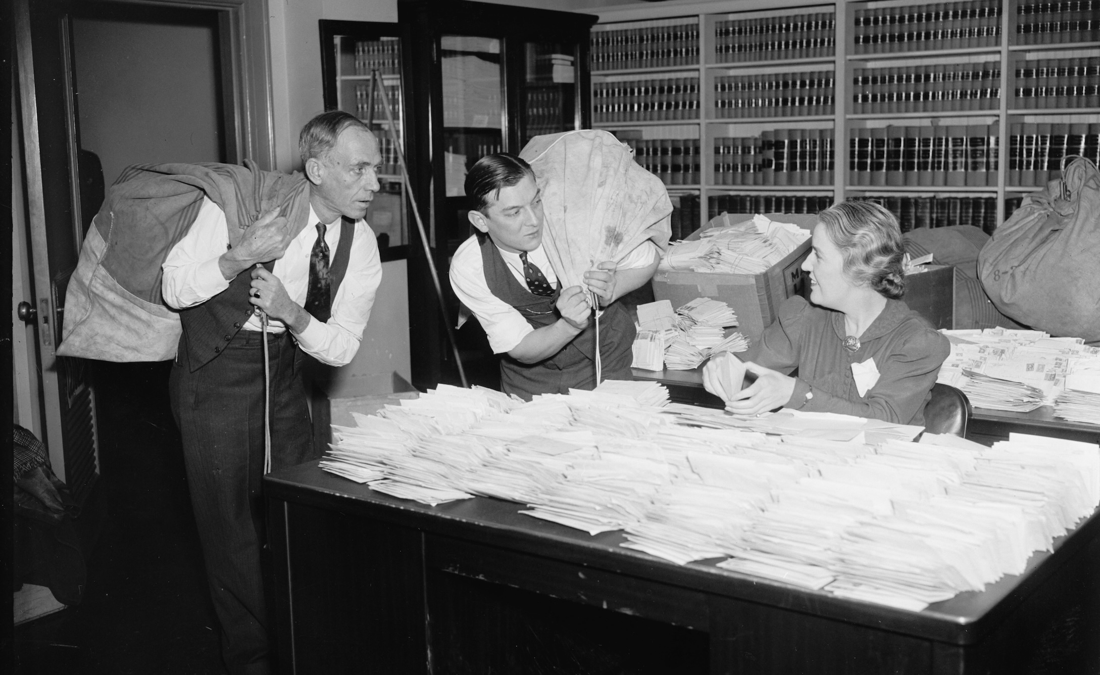 2 men carrying bags and woman seated at a desk opening letters