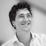 black-and-white photo of a man, smiling, in white shirt with glasses and earring