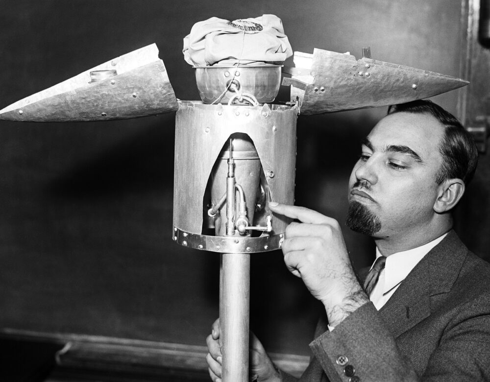 A black and white photograph of a man pointing at the open nose cone of a test rocket in a room with chalkboards on the walls