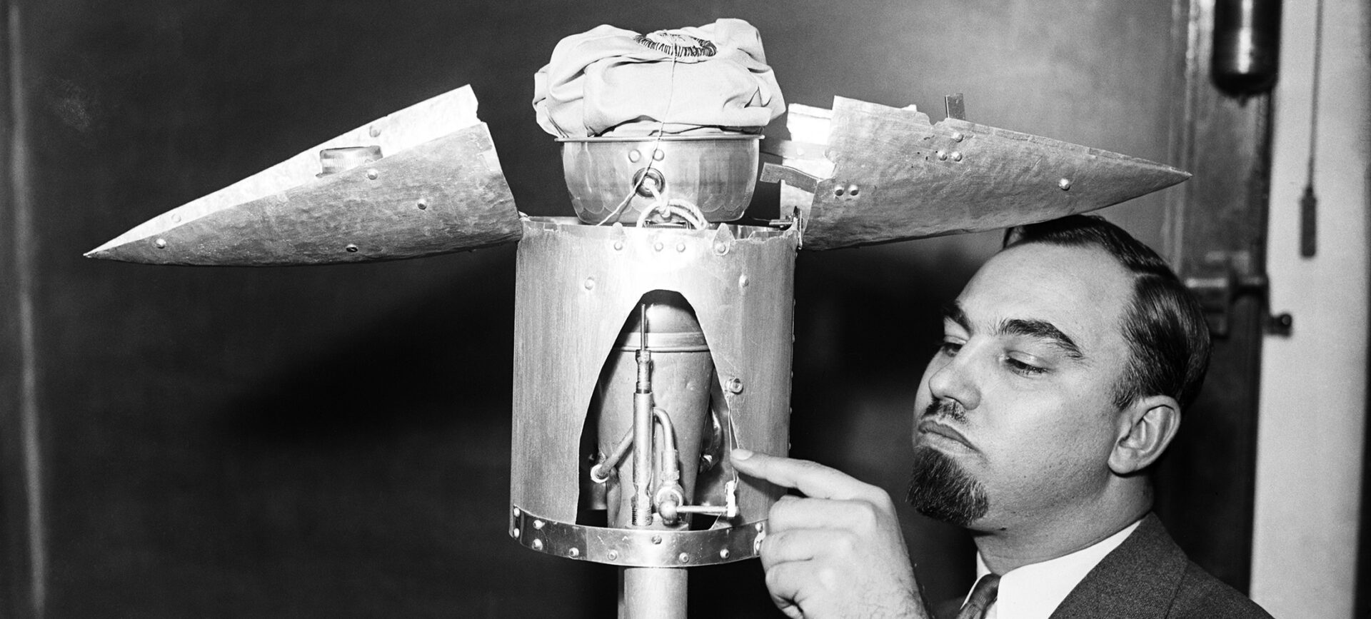 A black and white photograph of a man pointing at the open nose cone of a test rocket in a room with chalkboards on the walls
