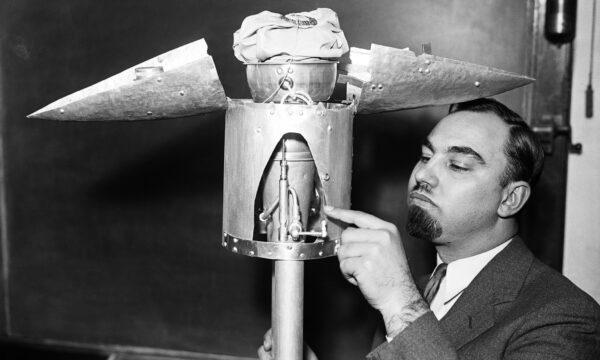 A black and white photograph of a man pointing at the open nose cone of a test rocket in a room with chalkboards on the walls