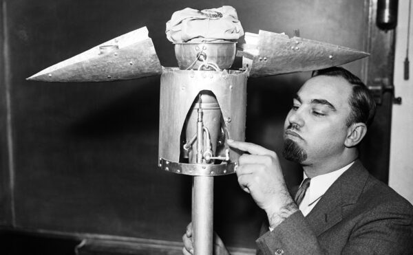 A black and white photograph of a man pointing at the open nose cone of a test rocket in a room with chalkboards on the walls