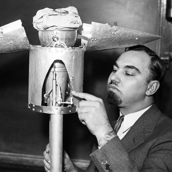 A black and white photograph of a man pointing at the open nose cone of a test rocket in a room with chalkboards on the walls