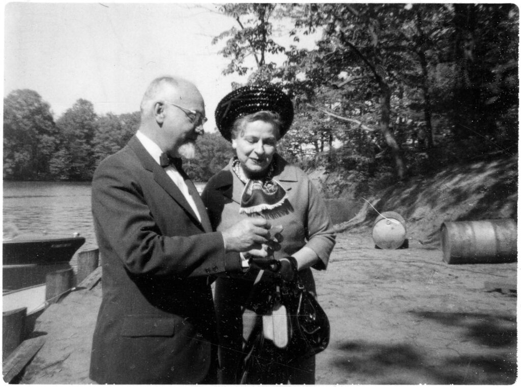A black and white photo of a man and woman looking at an unknown object on a beach, with trees and a lake in the background