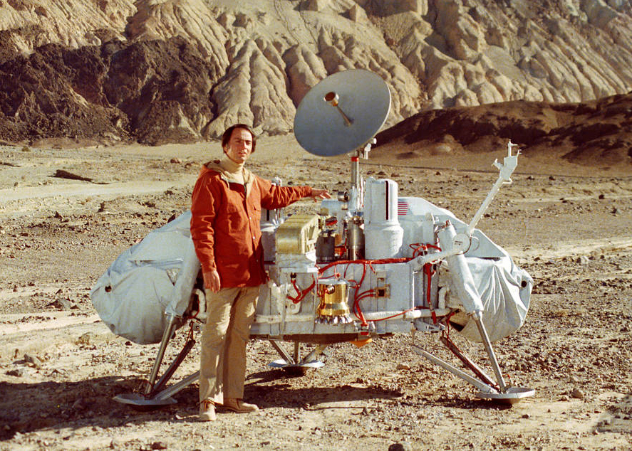 A color photo of a man standing next to a large space lander in a desert