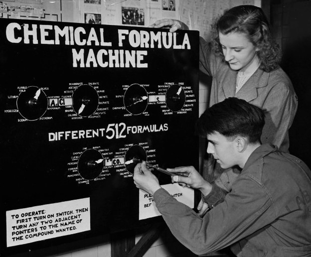 A black and white photo of a young man and woman operating a black, box-shaped device labeled “Chemical Formula Machine”