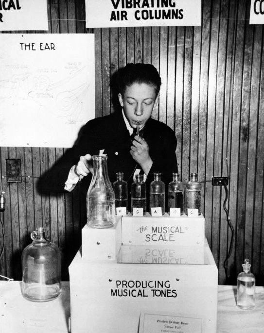 A black and white photo of a teenage boy blowing air through tubes to produce musical scales. A sign above him reads “Vibrating Air Columns”