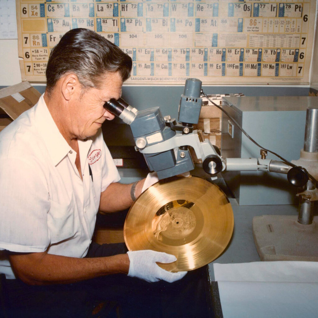A color photo of a man seated at a record plating machine and looking through a scope holding a golden record