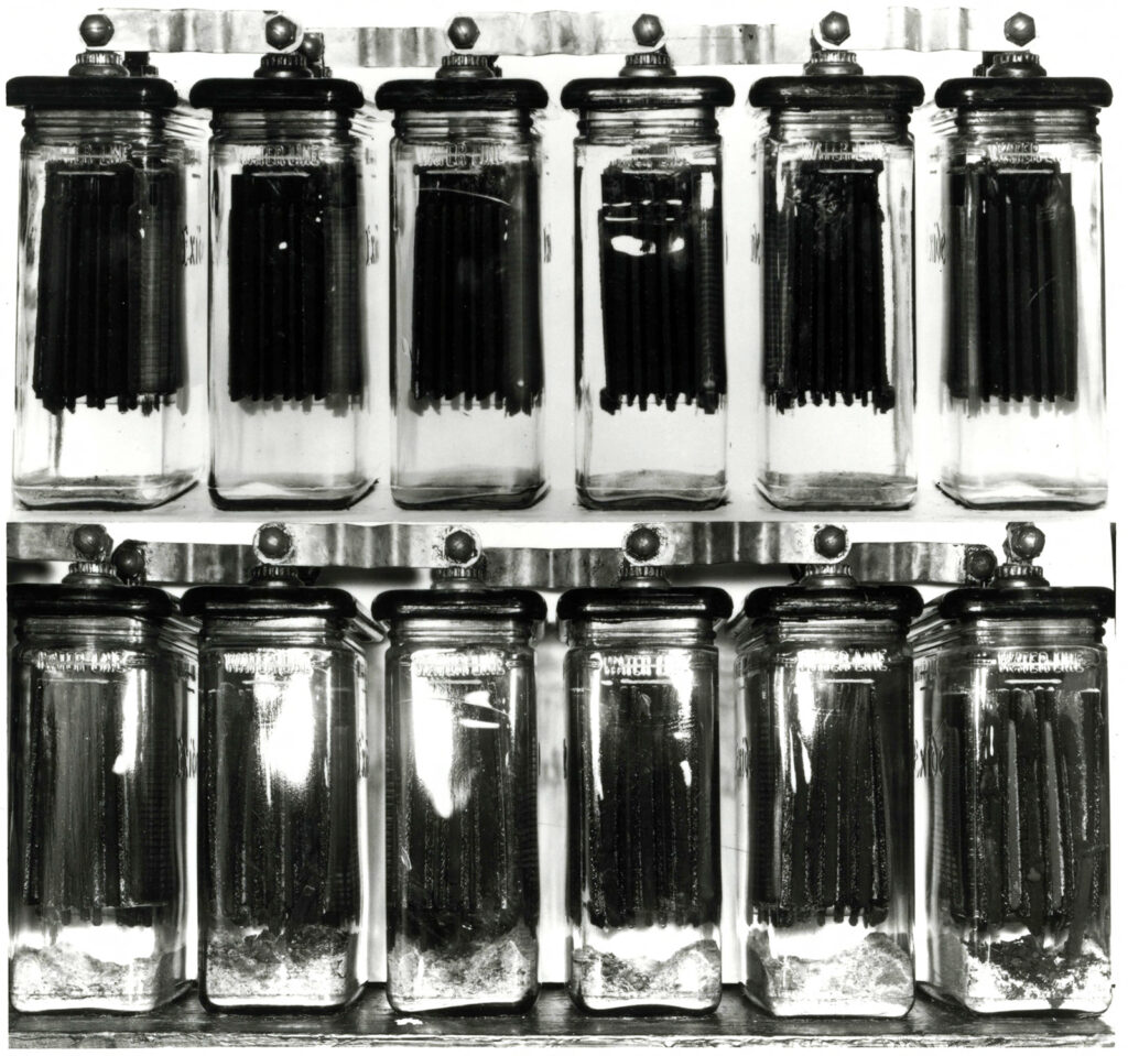 Black and white photo of two rows of six glass containers with car battery components inside them in a laboratory. “Water Line” is visible on the side of the containers.