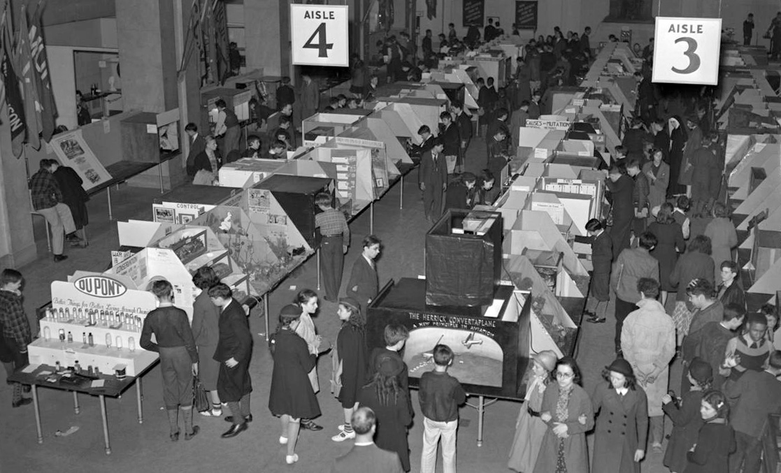 A black and white photo of a crowd of people observing exhibits and demonstrations at tables in a large room