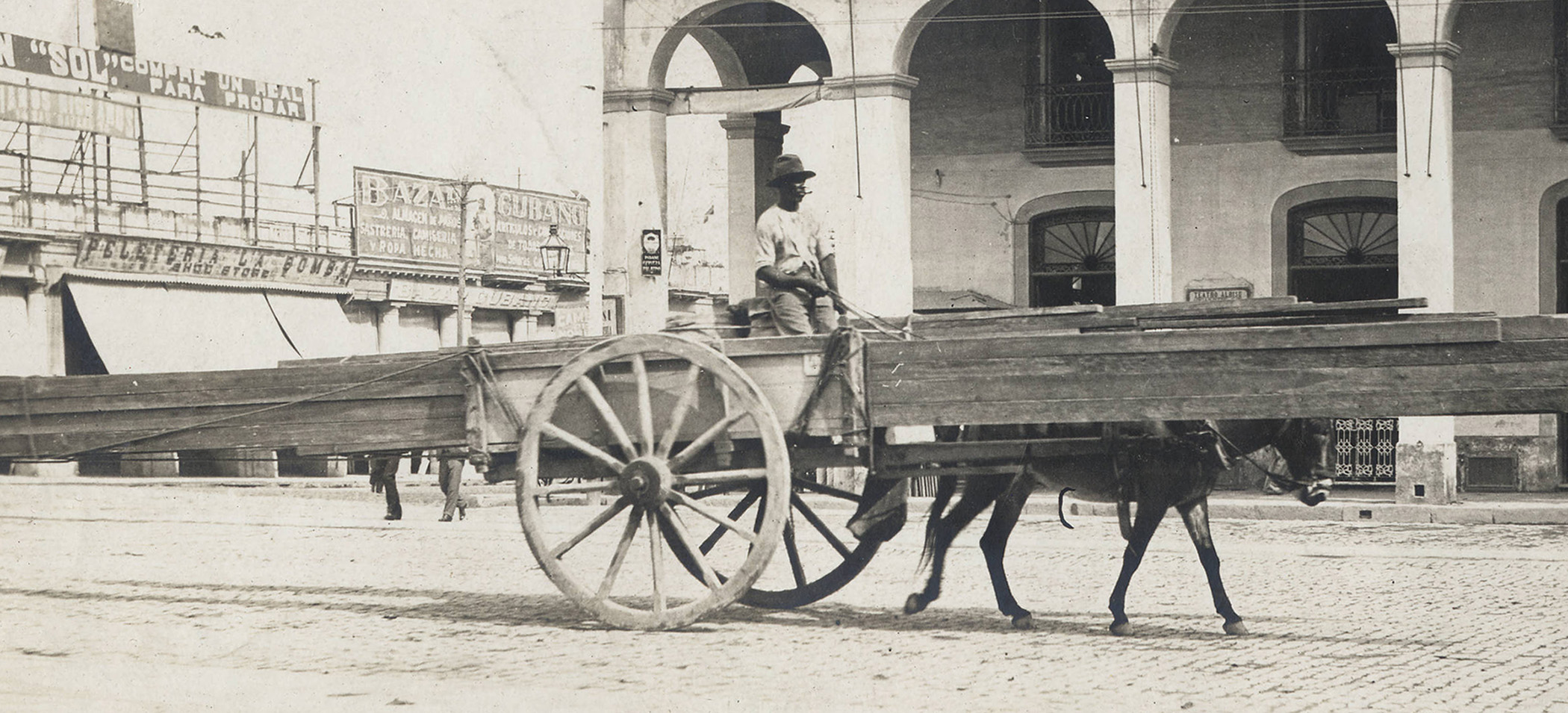 Foto antigua en blanco y negro de un hombre en una carreta cargada con una pila de largas piezas de madera en una calle de arquitectura colonial española; una sola mula tira de la carreta.