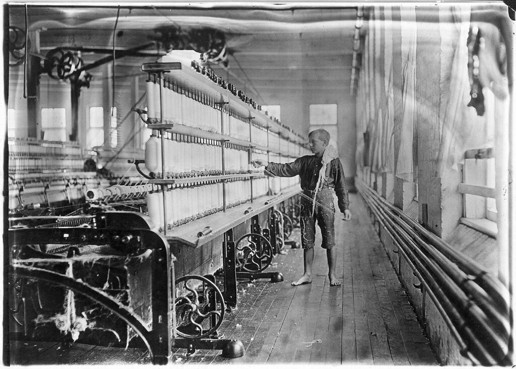 Foto en blanco y negro de un niño descalzo manipulando hilo conectado a una gran máquina de hilado en una fábrica.