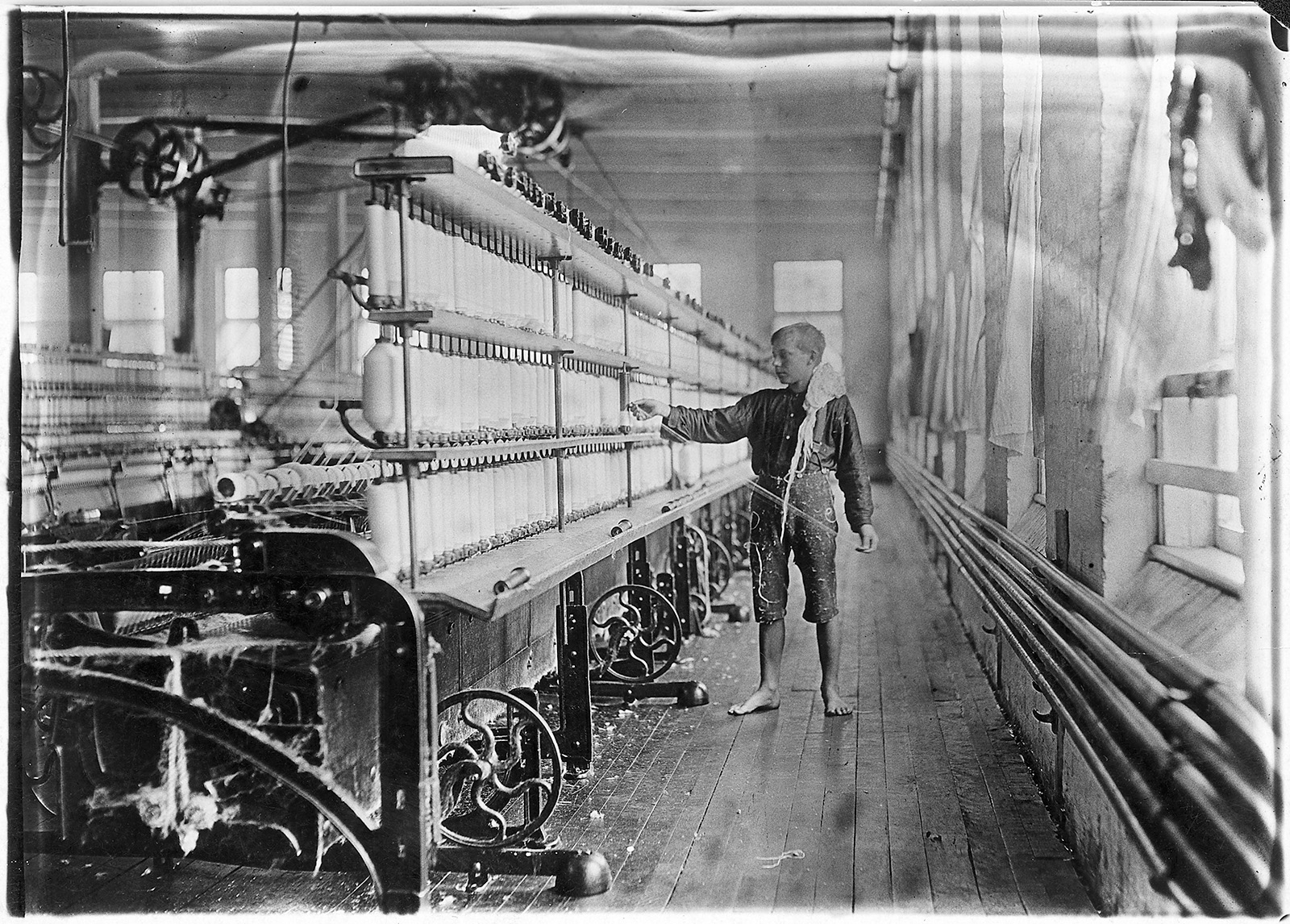 A black and white photo of a barefoot boy handling yarn attached to a larging spinning machine in a factory.