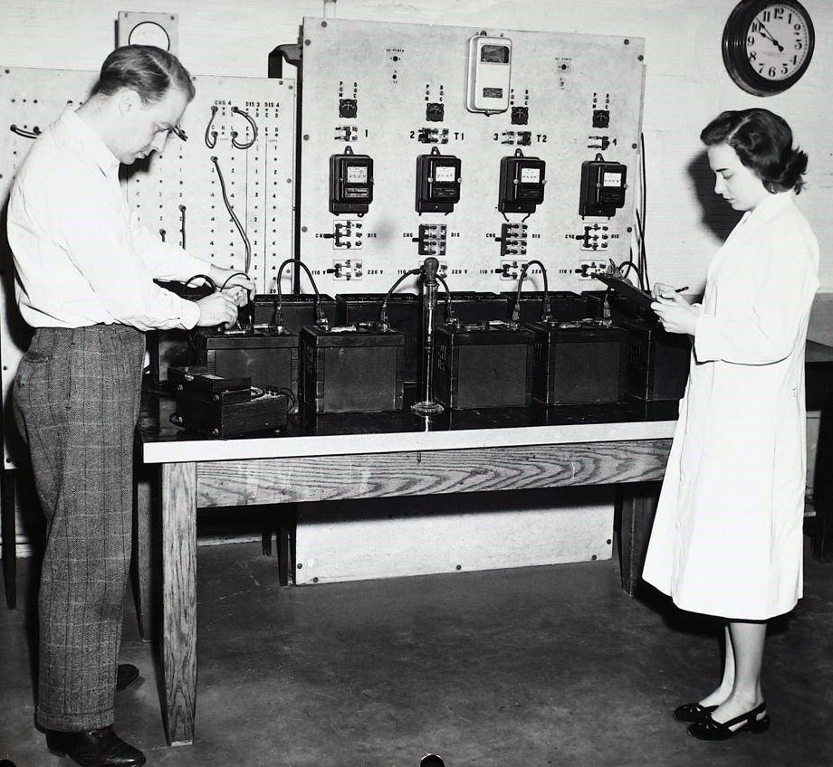 A black and white photo of a man and a woman testing automotive and aircraft batteries on a laboratory table. Behind the batteries, a panel of electrical equipment is attached to the wall.