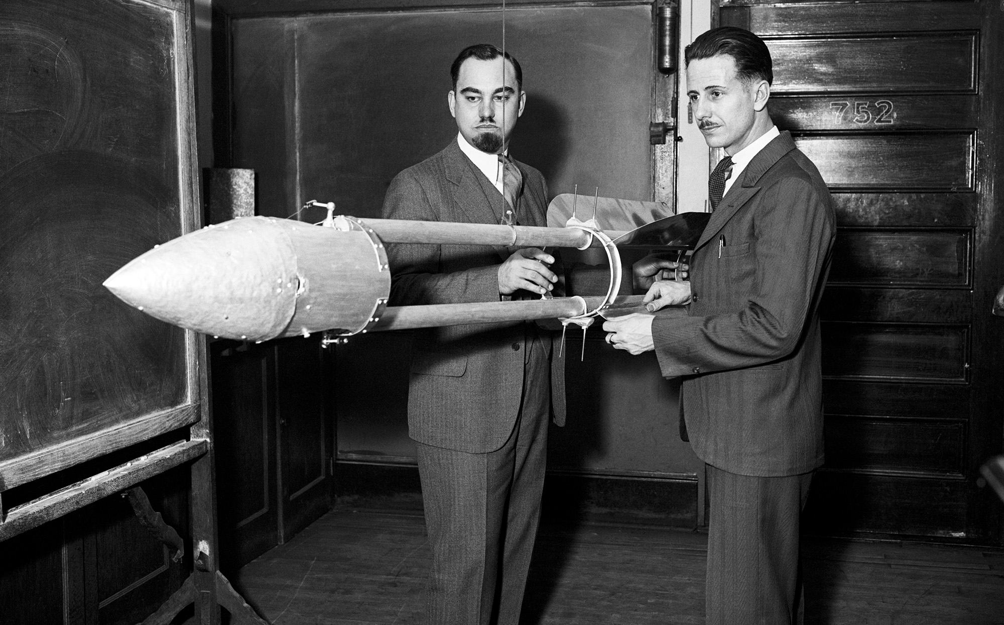 A black and white photo of two men holding a small rocket in a room surrounded by chalkboards