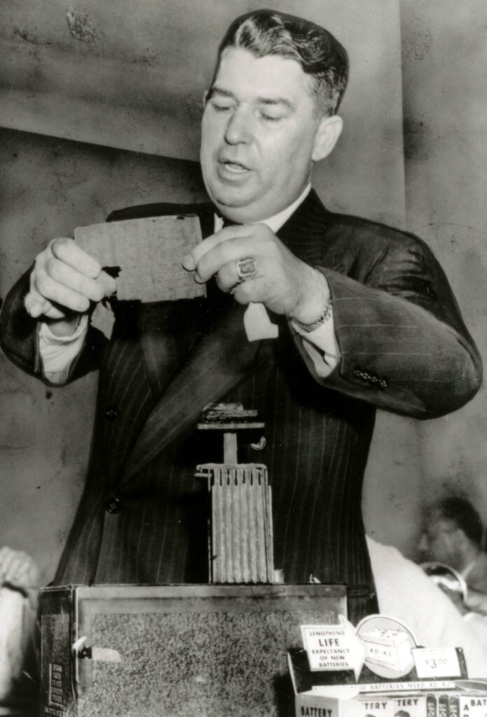 A black and white photo of a man in a suit holding up a worn car battery part, with a car battery on the table in front of him.