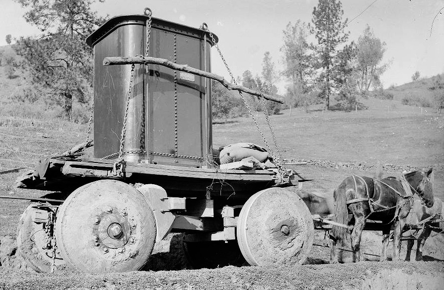 A black and white photo of two mules pulling a large wooden-wheeled cart with a rectangular metal object on top of it. Rocky hills and trees are in the background.