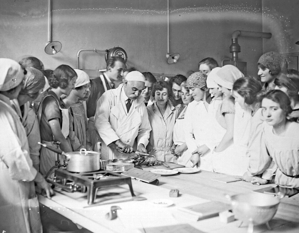 Black and white photograph of a man cutting food on a kitchen table surrounded by students